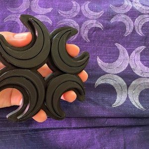 May include: A black, four-part crescent moon-shaped stamp held in a hand. The stamp is made of a dark material and has a layered design. The background is a purple fabric with a repeating pattern of the same crescent moon design.