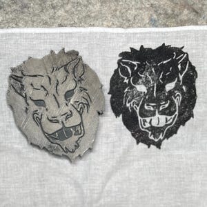 May include: Two lion head designs. One is a silver-colored metal, the other is a black print. Both have a detailed, stylized design of a lion's face with a smiling expression. The objects are on a white fabric surface.