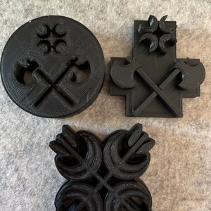 May include: Three black 3D-printed cookie cutters in different shapes. One is round with crossed axes and a decorative top. Another is cross-shaped with axes. The third is a four-leaf clover shape.