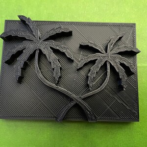 May include: A black rectangular decorative piece featuring two stylized palm trees. The palm trees are intertwined and raised above the surface, creating a 3D effect. The background has a textured, diagonal pattern.