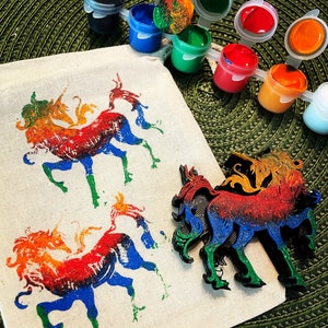 May include: A craft kit featuring a canvas bag with rainbow unicorn prints, a wooden unicorn cutout, and small paint pots in various colors. The unicorn design is in red, orange, yellow, green, blue, and purple.