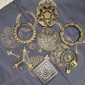 May include: A collection of black and gold decorative emblems, including a floral design, a laurel wreath, a pelican, and a chain-linked circle. The emblems are arranged on a dark gray fabric with gold accents.