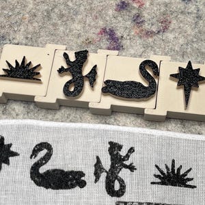May include: A set of four wooden blocks with black textured designs of a sunburst, lizard, swan, and star. Below, a fabric sample displays the same designs in black ink. The blocks are cream-colored.