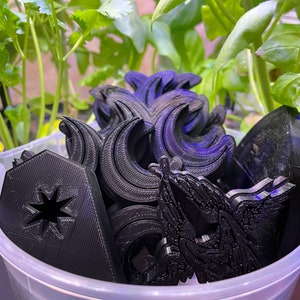 May include: A collection of black 3D-printed objects in a clear plastic container. The objects include crescent moons, a house shape with a star cutout, and other decorative shapes. Green plants are visible in the background.