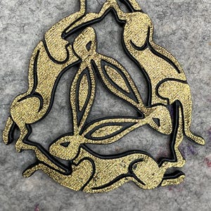 May include: A decorative wooden piece featuring a circular design of three hares. The hares are depicted in a running pose, with gold glitter filling and black outlines. The artwork is set against a gray background.