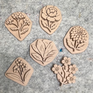 May include: A set of five light tan, flat, irregularly shaped stamps featuring raised floral designs. Each stamp displays a different botanical motif, including a cluster of small buds, a large blooming flower, a daisy-like bloom, and leafy stems.