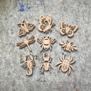 May include: A collection of nine light-colored wooden insect and butterfly cutouts arranged in a grid. The set includes various species such as a moth, butterflies, a dragonfly, a bee, a scorpion, a beetle, and a spider, all featuring detailed laser-cut patterns.