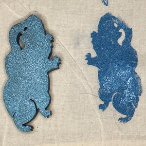May include: Two blue glittery cut-out figures of a stylized animal, possibly a squirrel or a similar rodent, are displayed on a white fabric background. The figure on the left has a textured surface, while the one on the right is a blue print.