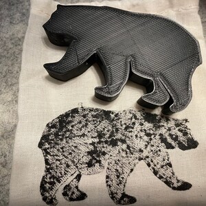 May include: A black bear-shaped object with a textured surface rests on a white cloth bag. The bag features a black and white bear illustration. The bear object appears to be made of plastic or a similar material.