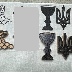 May include: A collection of wooden stamps featuring black ink designs of a dragon head, a chalice, a trident emblem, and a pitchfork. The set includes both flat printed outlines and three-dimensional wooden blocks with textured black surfaces.