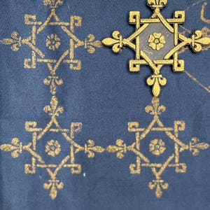 May include: Close-up of a navy blue fabric with a repeating gold pattern. The pattern includes fleur-de-lis and geometric shapes. A gold decorative element with a floral center is on the fabric.