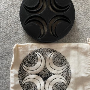 May include: A black, circular object with four crescent moon shapes cut out, next to a white canvas bag with a matching design. The bag features a black and white speckled background with four white crescent moon shapes.