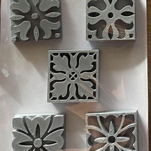 May include: Five gray, square decorative tiles with floral patterns. Each tile features a different variation of the floral design, with openwork details. The tiles are arranged on a white surface, possibly for display or sale.