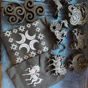 May include: An assortment of black decorative items, including Celtic symbols, mythical creature-shaped coasters, and drawstring bags with celestial and mythological designs. The items are arranged on a blue surface.