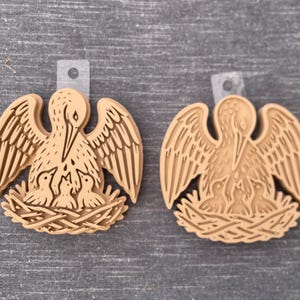 May include: Two tan, circular, molded plastic ornaments featuring a raised relief design of a pelican with wings spread wide, tending to three small chicks nestled within a woven nest. Each ornament includes a clear plastic tab with a hole.