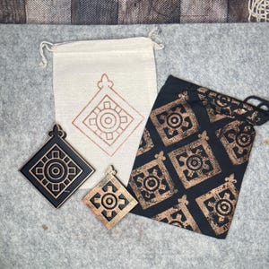 May include: A collection of decorative items featuring a geometric design. Includes two drawstring bags, one cream-colored and one black, and two square ornaments. The ornaments are black and gold-colored, and the bags have a repeating pattern.