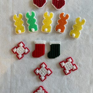 May include: A collection of colorful, cross-stitched ornaments. The assortment includes red hearts, bunny rabbits in yellow, green, and orange, red and green stockings, and red and white snowflake designs. The ornaments are likely handmade and suitable for holiday decor.