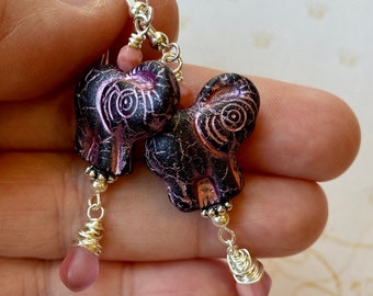 May include: A pair of dangle earrings featuring a purple and black elephant charm with silver accents and a pink glass bead on each earring.