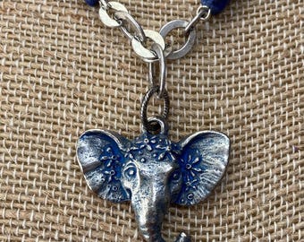 May include: Silver-toned elephant pendant necklace with blue accents. The elephant pendant is detailed with floral designs and hangs from a silver chain. The necklace features blue stone beads.