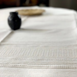 Puede incluir: Primer plano de un mantel blanco con detalles texturizados y el texto "OAXACA BY HAND" en gris. Un pequeño jarrón negro y un plato tejido están ligeramente desenfocados en el fondo. El mantel tiene un diseño sutil y elegante.