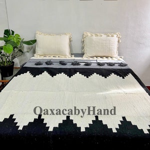 K&ouml;nnte beinhalten: Eine gewebte Decke mit einem geometrischen Schwarz-Wei&szlig;-Muster, grauen Akzenten und Quasten. Zwei cremefarbene Kissen mit Fransen liegen darauf. Der Text "OaxacabyHand" ist auf der Decke zu sehen.