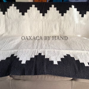 K&ouml;nnte beinhalten: Eine schwarz-wei&szlig;e gewebte Decke mit geometrischem Muster. Die Decke ist &uuml;ber ein Sofa drapiert. Der Text "OAXACA BY HAND" ist auf die Decke gedruckt.