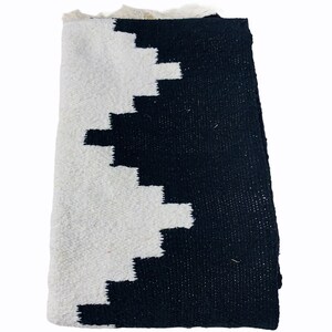 K&ouml;nnte beinhalten: Schwarz-wei&szlig; gewebter Stoff mit geometrischem Muster. Das Design zeigt ein Zickzackmuster in Wei&szlig; auf schwarzem Hintergrund. Der Text "OAXACA BY HAND" wird unten angezeigt. Der Stoff hat einen Fransenrand.