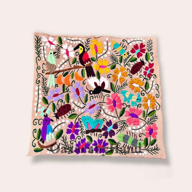 K&ouml;nnte beinhalten: Quadratischer, bestickter Textilartikel mit einem lebendigen Blumen- und Fauna-Design. Das Kunstwerk zeigt farbenfrohe V&ouml;gel, Blumen und Insekten in Rosa, Lila, Gelb und Gr&uuml;n auf beigem Hintergrund. Der Textilartikel ist wahrscheinlich handgefertigt.