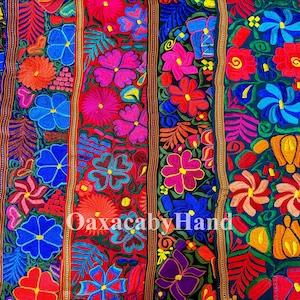 Puede incluir: Un textil con un denso bordado floral de colores en tonos rosa, azul, naranja y morado sobre un fondo oscuro. La tela está dividida en secciones verticales por bandas tejidas decorativas. El texto OaxacabyHand es visible.