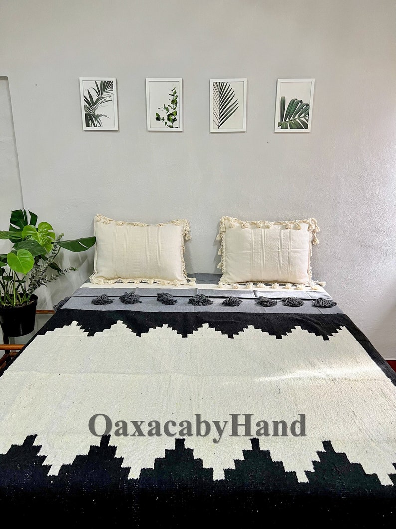 K&ouml;nnte beinhalten: Eine gewebte Decke mit einem geometrischen Schwarz-Wei&szlig;-Muster, grauen Akzenten und Quasten. Zwei cremefarbene Kissen mit Fransen liegen darauf. Der Text "OaxacabyHand" ist auf der Decke zu sehen.