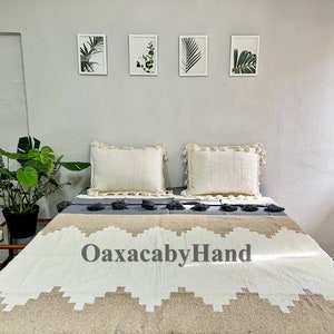 K&ouml;nnte beinhalten: Ein Bett mit einer cremefarbenen Decke mit einem geometrischen Design in Beige und Wei&szlig;. Zwei cremefarbene Kissen mit Fransenbesatz liegen auf dem Bett. Der Text "OaxacabyHand" ist auf der Decke zu sehen. Gerahmte botanische Drucke h&auml;ngen an der Wand.