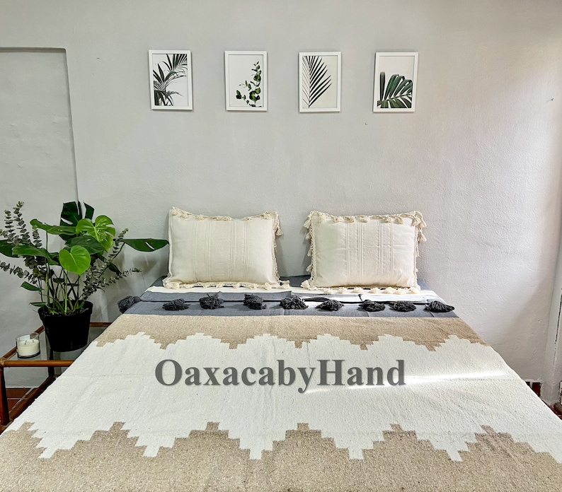 K&ouml;nnte beinhalten: Ein Bett mit einer cremefarbenen Decke mit einem geometrischen Design in Beige und Wei&szlig;. Zwei cremefarbene Kissen mit Fransenbesatz liegen auf dem Bett. Der Text "OaxacabyHand" ist auf der Decke zu sehen. Gerahmte botanische Drucke h&auml;ngen an der Wand.