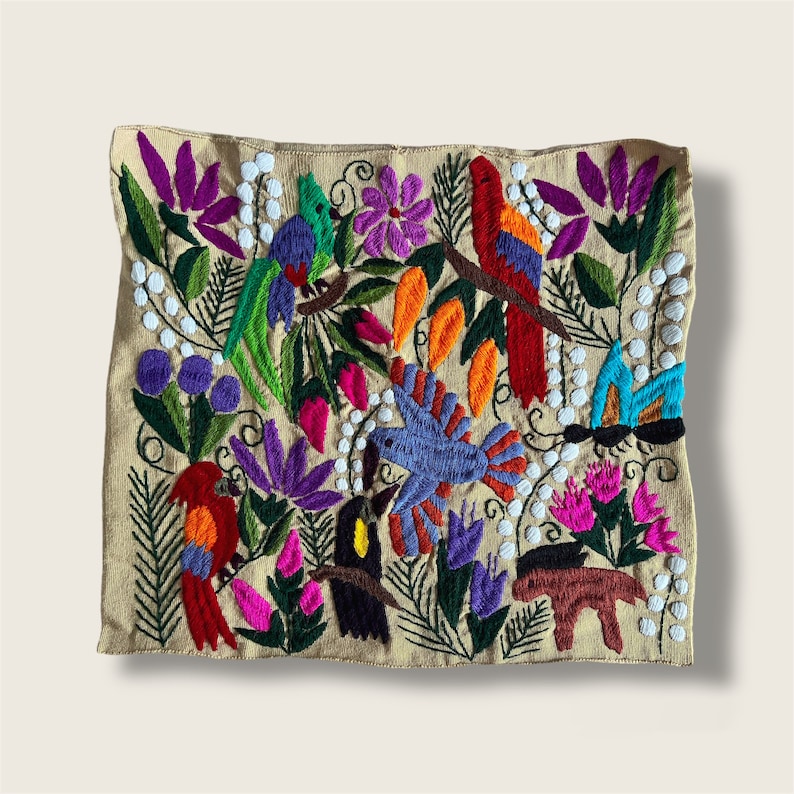 K&ouml;nnte beinhalten: Quadratischer, beigefarbener Kissenbezug mit farbenfrohen Stickereien von V&ouml;geln, Blumen und Bl&auml;ttern. Das Design zeigt Papageien, Schmetterlinge und andere V&ouml;gel in Rot, Blau, Gr&uuml;n und Lila. Der Kissenbezug hat ein detailliertes, handwerkliches Aussehen.
