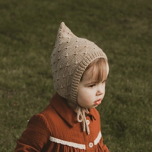 May include: A child in a rust-coloured dress with white lace trim and a matching cape. They are wearing a knitted bonnet and brown leather boots. The child is walking on green grass.