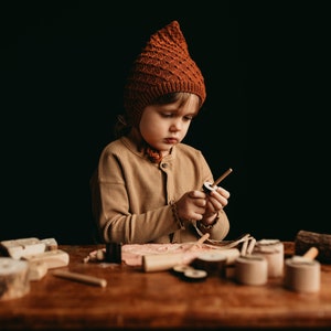 以下が含まれることがあります： 茶色のニット帽とジャケットを着た子供が、木製のテーブルで木製のおもちゃを組み立てることに集中しています。テーブルには、円柱、円盤、ダボなど、さまざまな木製のおもちゃのパーツが置かれています。背景は暗い色です。