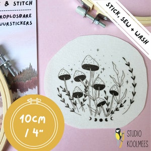 Op de afbeelding: Een witte viltcirkel met een zwarte lijntekening van paddenstoelen en bladeren. De cirkel heeft een diameter van 10 cm en heeft de tekst "STICK, SEW + WASH" in de rechterbovenhoek.