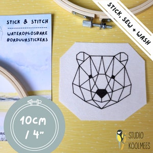 May include: A geometric bear embroidery design on a white fabric, measuring 4 inches (10cm) in diameter. The design is accompanied by a wooden embroidery hoop and a package labeled "Stick & Stitch". The text "Stick, Sew + Wash" is also visible.