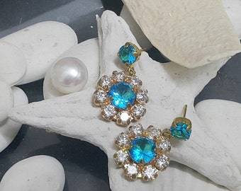 Puede incluir: Un par de pendientes colgantes de oro con piedras de cristal azules y transparentes. Los pendientes tienen un diseño en forma de flor con una piedra de cristal azul más pequeña que cuelga del centro.