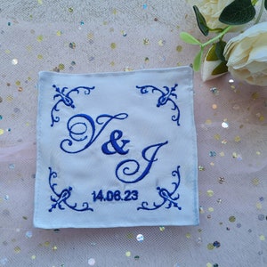 Può includere: Tessuto quadrato bianco con lettere ricamate in blu. Le lettere sono in un font corsivo e dicono "V & J" con la data "14.06.23" sotto.