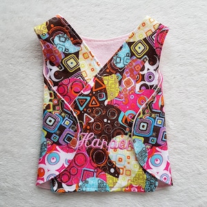 Puede incluir: Un babero para bebé estampado con interior rosa. El babero presenta un diseño vibrante de formas geométricas en rosa, marrón, azul y naranja. El nombre "Harper" está bordado con hilo rosa en la parte delantera.