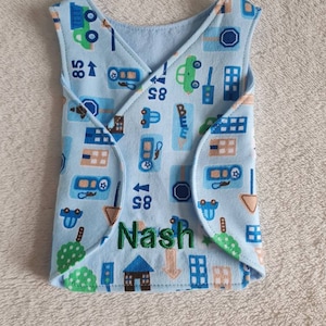 Puede incluir: Un babero para bebé azul claro con un estampado de ciudad con coches, casas y señales de tráfico. El nombre "Nash" está bordado en verde. El babero tiene un diseño cruzado.