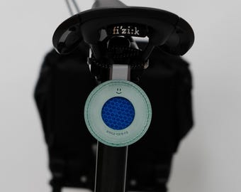 Può includere: Sella per bicicletta con il marchio "fi'zi:k". Un accessorio rotondo verde chiaro con un motivo a nido d'ape blu e una faccina sorridente è attaccato al reggisella. L'accessorio ha testo in lingua straniera.