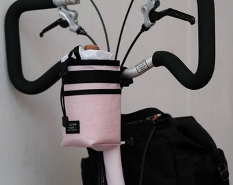 May include: A light pink cylindrical bicycle handlebar bag with black trim and a drawstring closure. The bag features a small black rectangular label with the white text PRACTICO ARTE CRAFT & DESIGN. It is mounted on the handlebars of a bicycle.