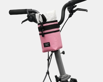 May include: A pink bicycle handlebar bag with black detailing and a white interior. The bag is mounted on the handlebars of a bicycle. The bag features a small rectangular label with text. The handlebars are black.