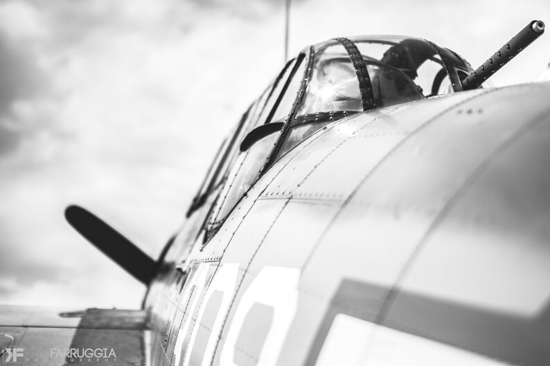 May include: A black and white photo of a vintage airplane cockpit. The cockpit is open and the pilot's seat is visible. The airplane is painted in a silver color with a black propeller.