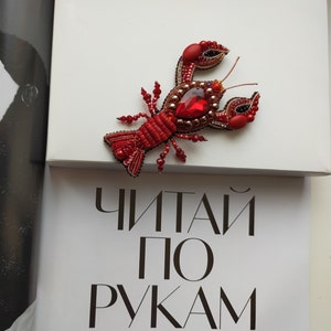 May include: A red beaded lobster brooch with a large red crystal in the center. The brooch is sitting on a white surface with the text "ЧИТАЙ ПО РУКАМ" in black.