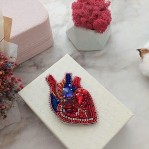 May include: A red, blue, and gold beaded heart-shaped brooch with rhinestones and sequins. The brooch is on a white box with a textured surface.