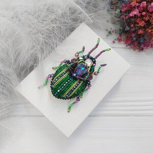 Peut inclure: Une broche en forme de scarab&eacute;e en perles vertes et violettes avec un corps orn&eacute; de bijoux. La broche est sur une surface blanche.