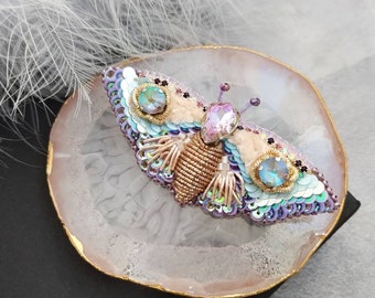 May include: A beaded butterfly brooch with iridescent sequins, pink and blue crystals, and gold accents. The brooch is displayed on a pink and white agate slice.