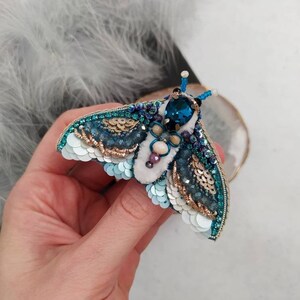 May include: A blue and white beaded moth brooch with iridescent sequins and a large blue gemstone in the center.